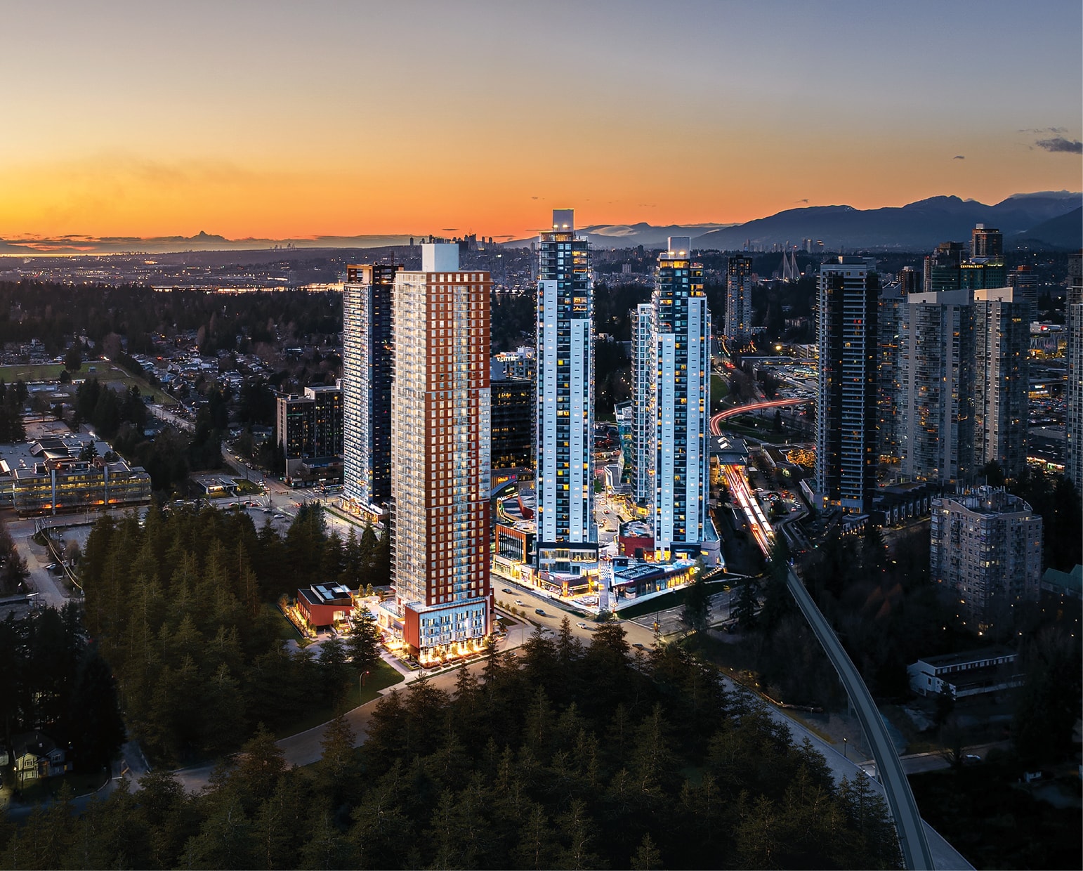 High-rise city skyline at sunset with mountains, roads, trees, and illuminated buildings.
